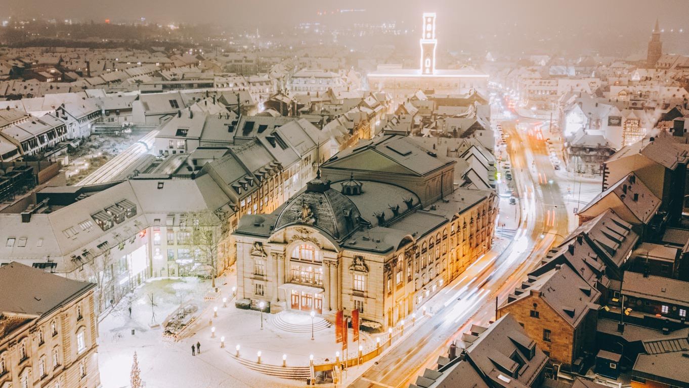 Blick von oben auf das verschneite Stadttheater am Abend. Im Hintergrund der beleuchtete Rathausturm.